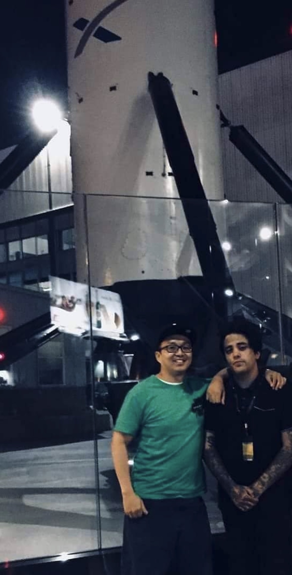 Ernest Yeung and Eros Marcello at SpaceX headquarters at night, beneath a recovered Falcon 9 first stage with its landing legs deployed.